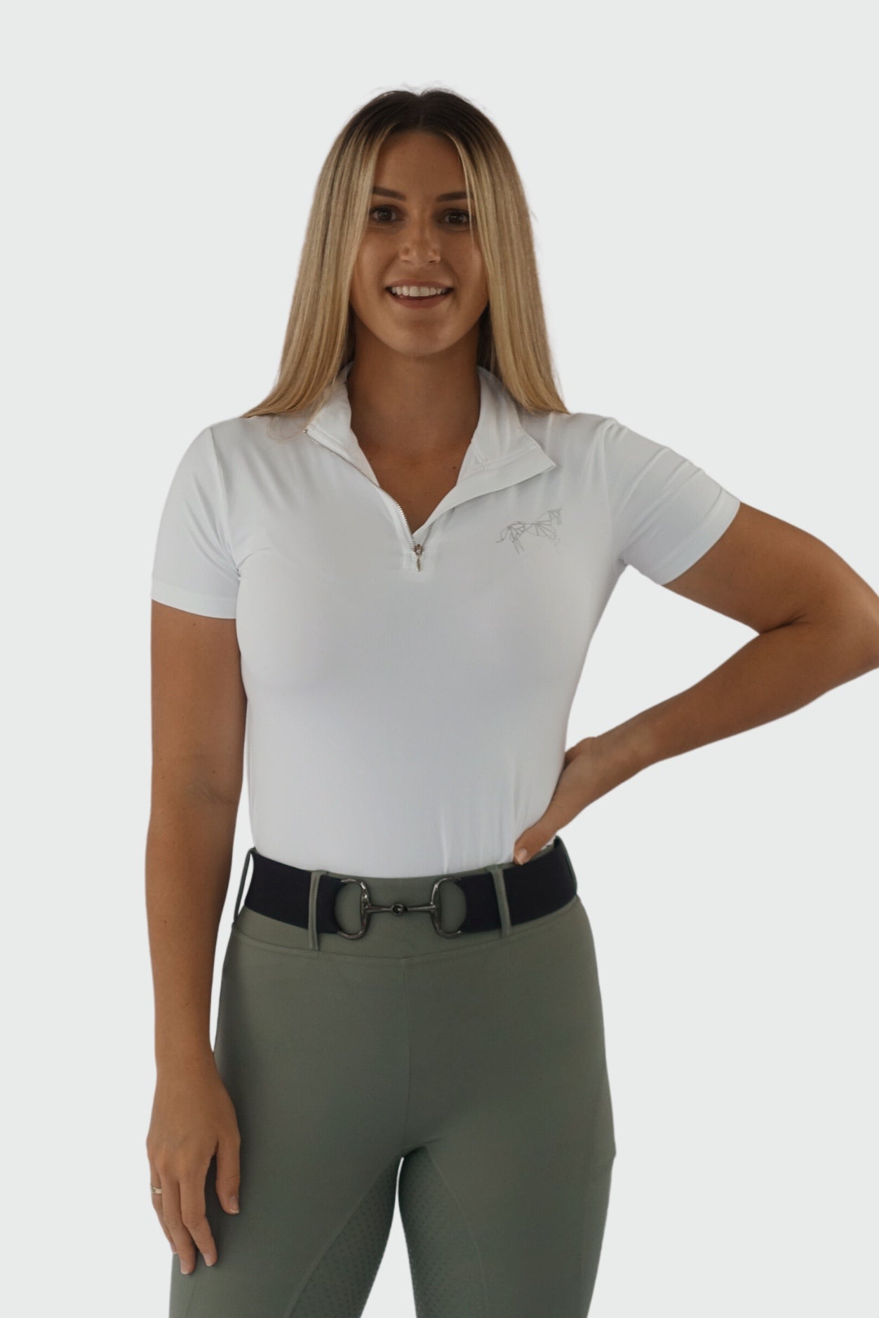 A person with long blonde hair stands confidently with a slight smile, wearing a fitted white Pure Canter V1 Motion Top Short and high-waisted green pants with a black belt that has a double-ring buckle. One hand is on their hip while the other rests by their side.