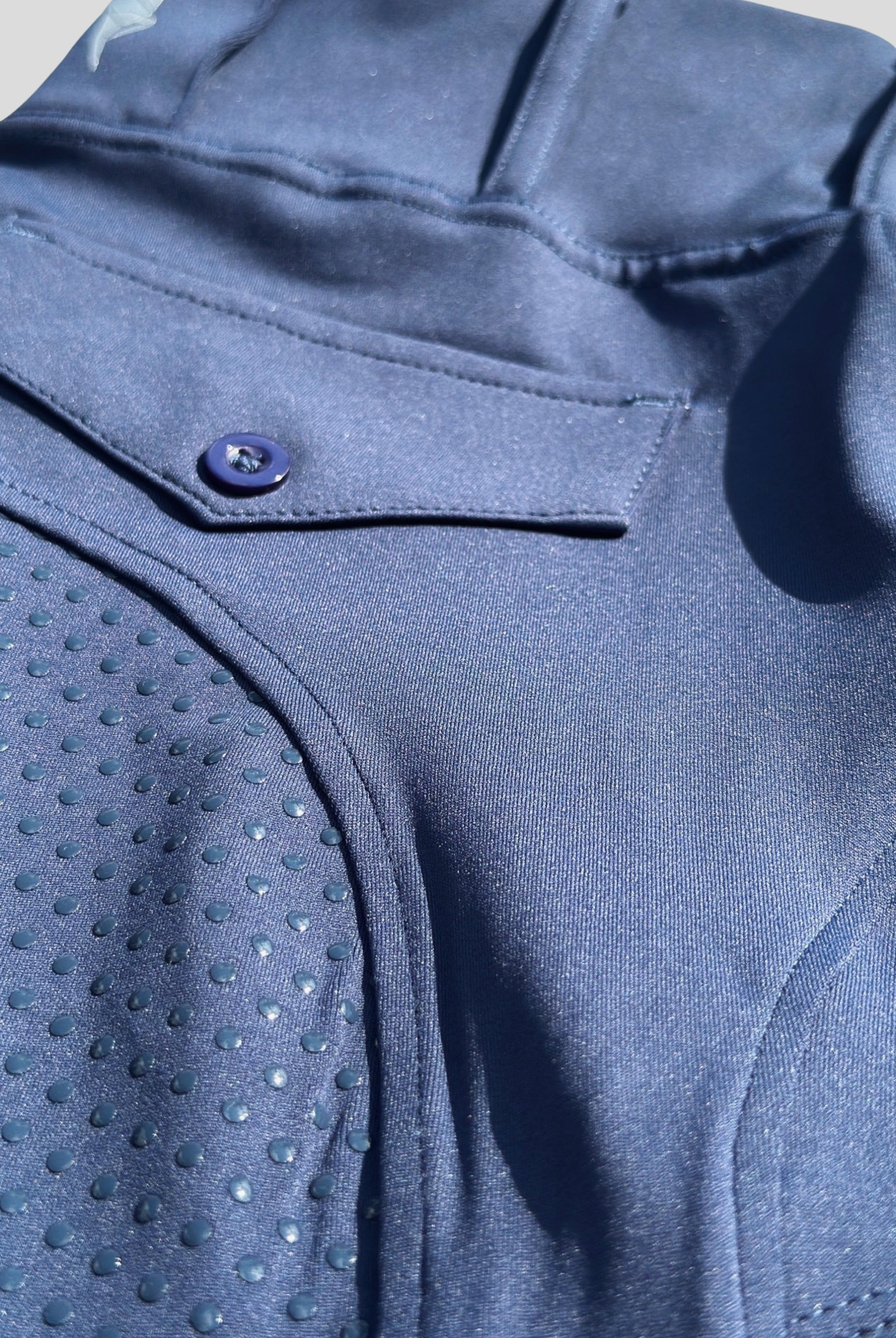 Close-up image of a section of a dark blue garment, likely V2 Fusion Riding Tights - Navy by Pure Canter. The fabric features a smooth texture and a dotted, textured pattern on one part. A visible button and pocket detail are also present, as well as stitching lines. The image is well-lit, showcasing equestrian fashion at its finest.