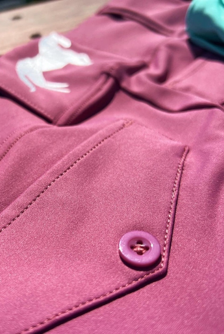 Close-up view of vibrant pink **V1 Fusion Riding Tights - Mulberry** with a button, pocket detail, and a white logo of a horse on the performance fabric by **Pure Canter**. A light blue object is slightly visible in the background.