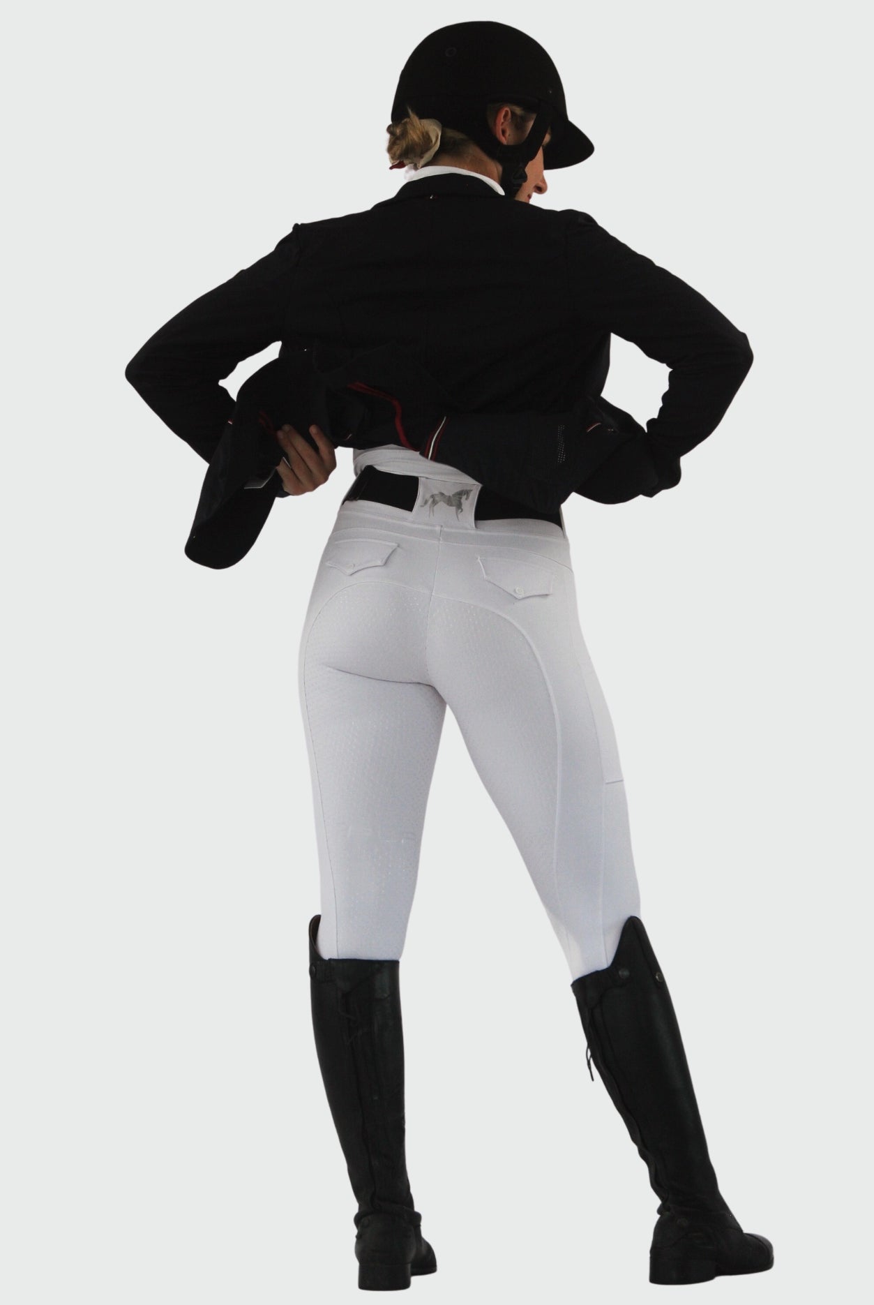 A person in equestrian attire stands with their back to the camera. They are wearing a black riding helmet, black jacket, Pure Canter Competition Tights - White, and black riding boots. The person appears to be adjusting their jacket made from performance fabric. The background is plain white.