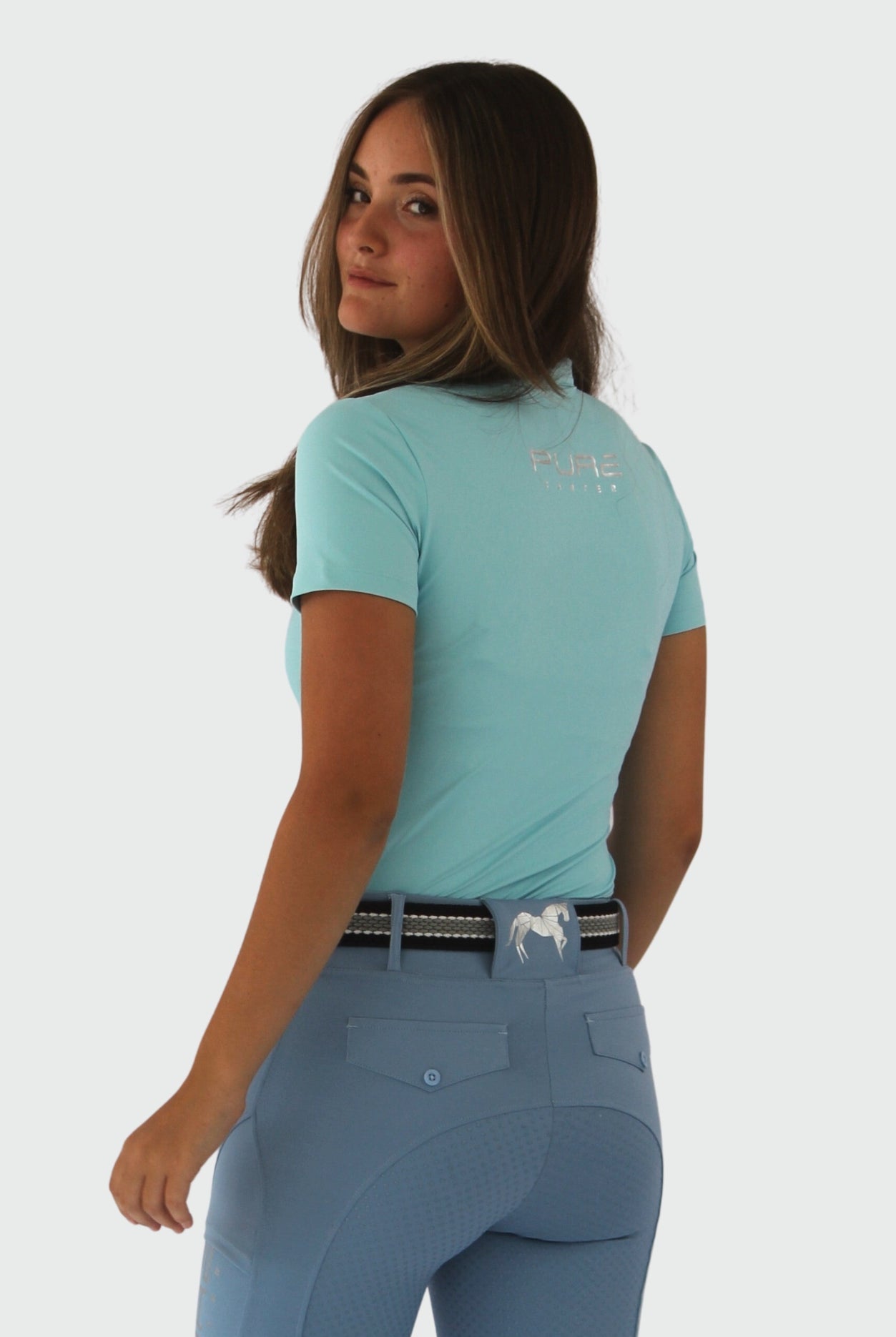 A person with long hair is standing and looking over their shoulder towards the camera. They are wearing the Pure Canter V1 Motion Top Short - Ocean Blue made from breathable fabric and matching blue pants with a silver belt and horse emblem on the back pocket.
