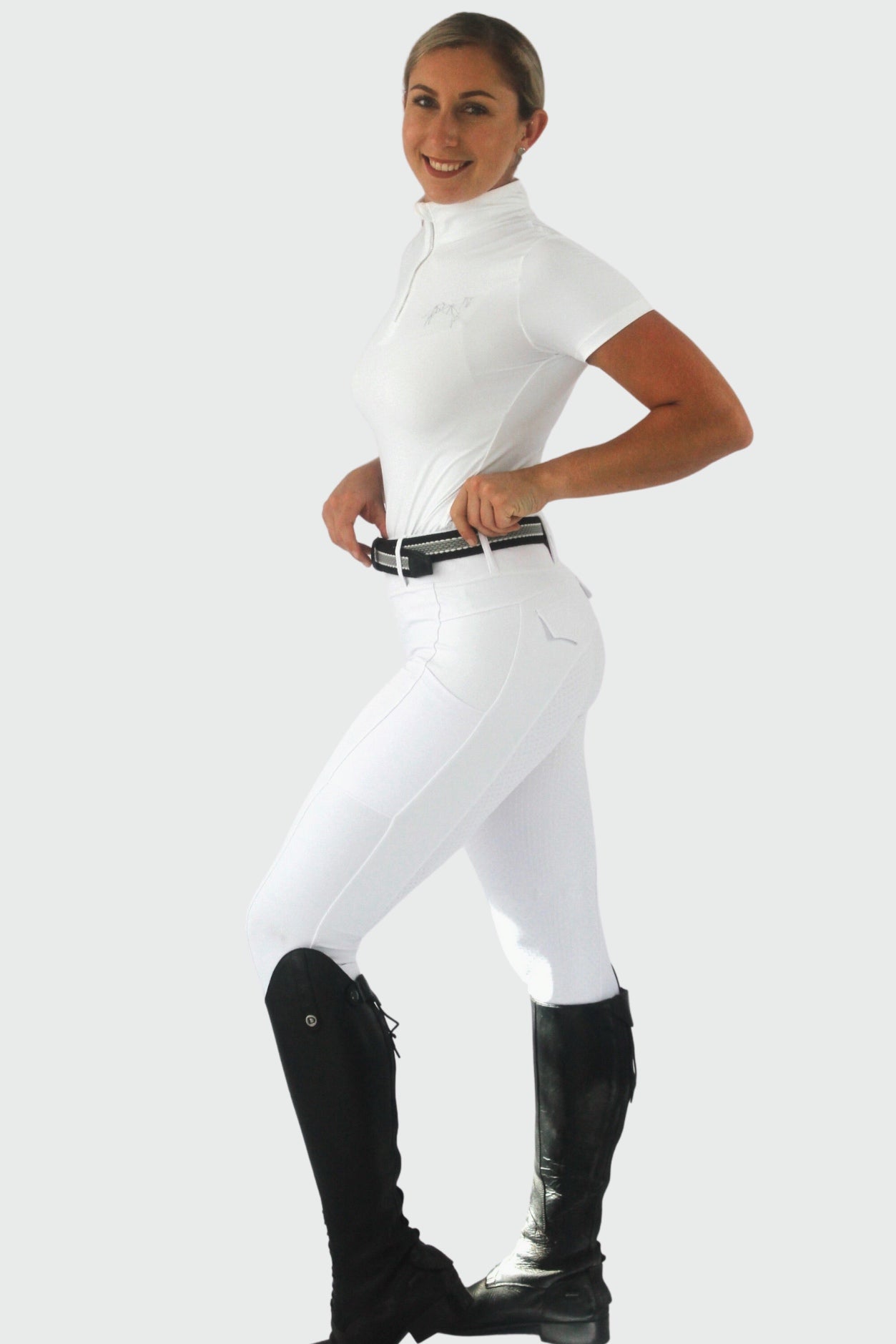 A person is posing and standing sideways, wearing a white short-sleeve riding shirt tucked into white Pure Canter Competition Tights made from performance fabric. The person has a black belt, black knee-high riding boots, and is smiling. The background is plain white.