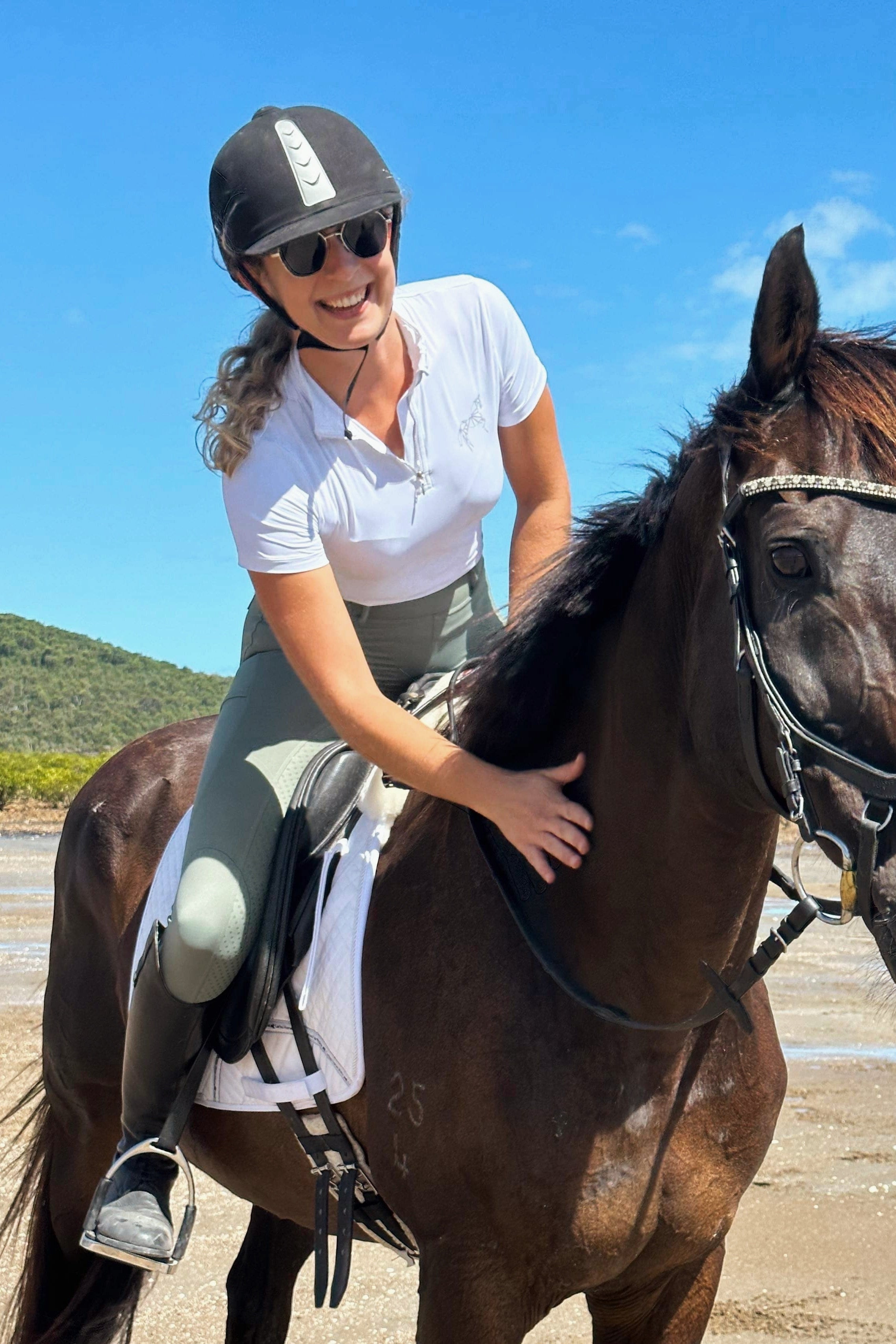 A smiling woman wearing a helmet, sunglasses, and a Pure Canter V1 Motion Top Short - White is sitting on a dark-colored horse. She is gently patting the horse's neck on a sunny day, with clear blue skies and greenery in the background. The scene appears to be on a beach.
