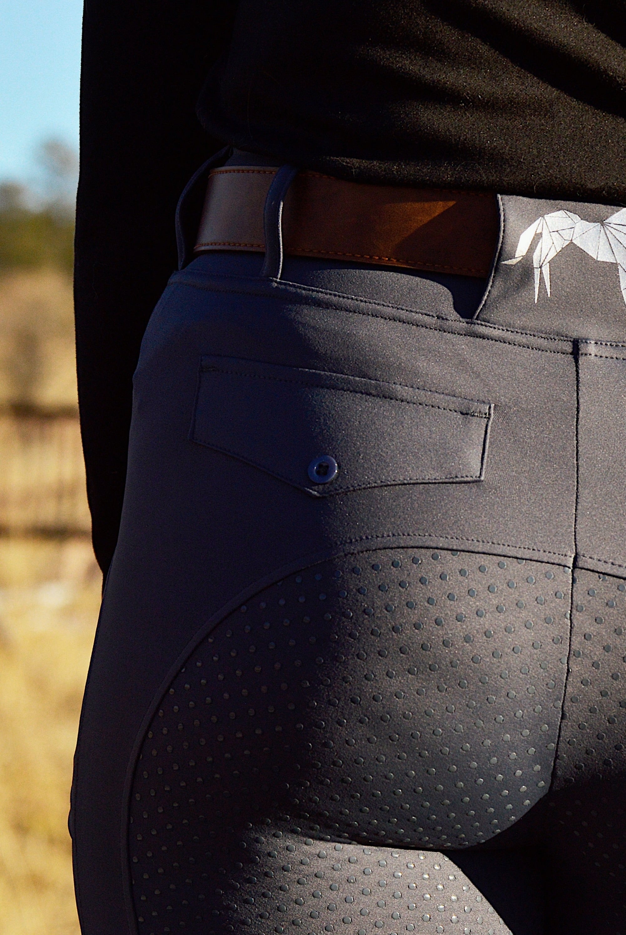 Close-up of a person from the waist down wearing form-fitting dark gray Pure Canter V2 Fusion Riding Tights - Dark Grey with small dots on the seat and upper thigh areas for grip, embodying equestrian fashion. They are also wearing a brown belt threaded through the belt loops and a black waistband from a shirt or jacket.