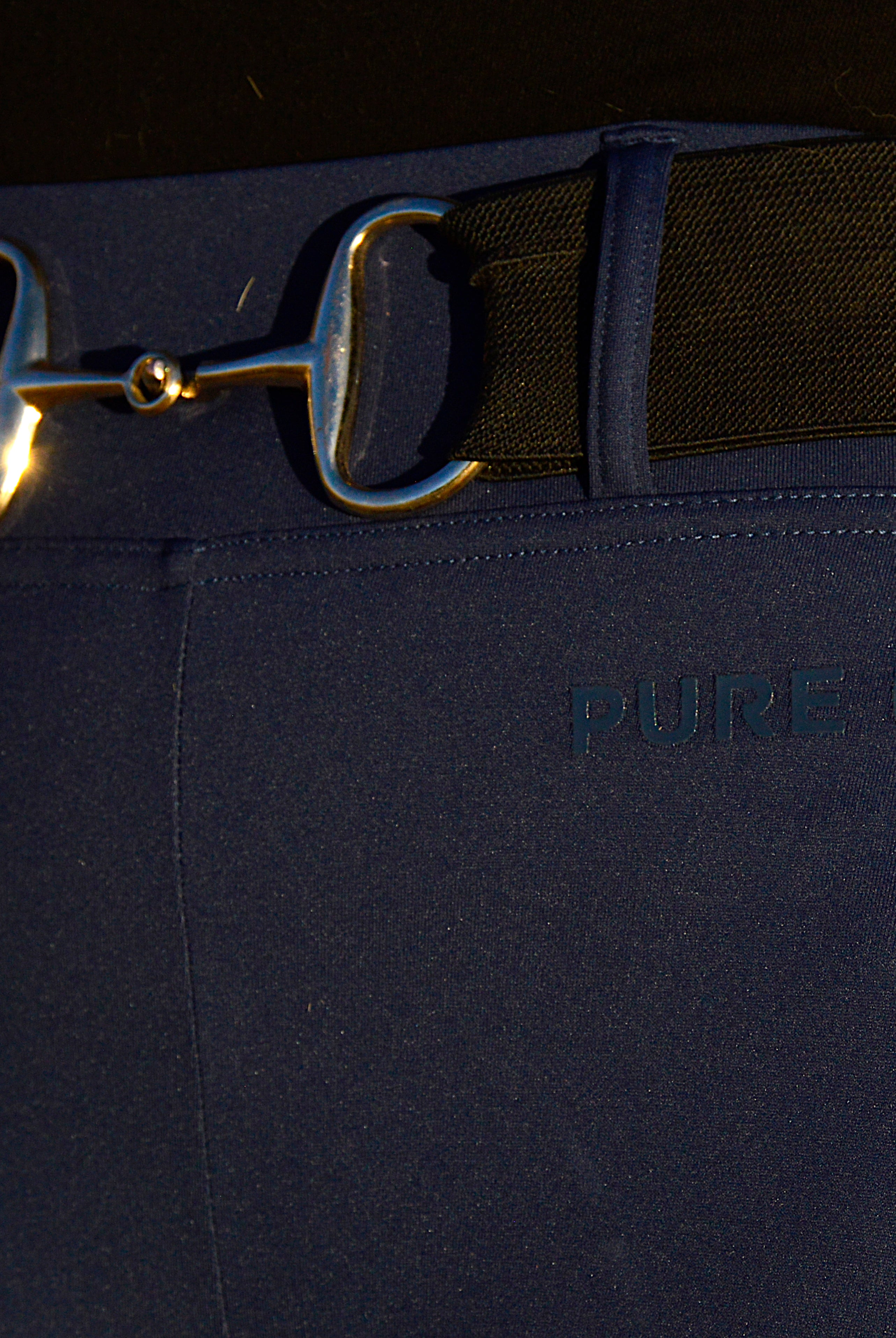 Close-up of a person wearing blue Fusion Tights G2 - Navy and a belt featuring a horse bit buckle. The waistband of the pants has "PURE CANTER" printed on it. The person is also wearing a red plaid shirt, showcasing an impeccable touch of equestrian fashion.