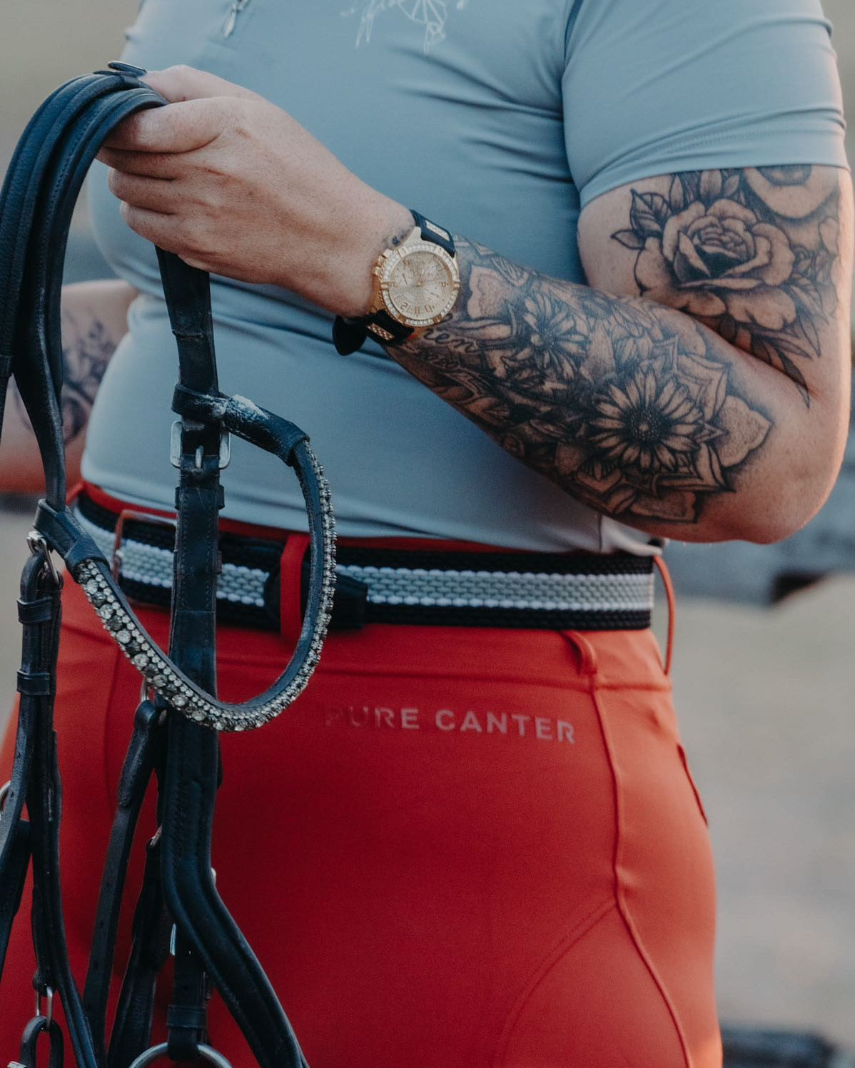 A person with tattooed arms wearing a blue shirt and bright red pants holds horse reins. The individual sports a gold watch on their left wrist. Blending equestrian fashion, the shirt features a subtle horse design while the pants, V2 Fusion Riding Tights - Outback from Pure Canter, are made of performance fabric and have "PURE CANTER" written on the hip.