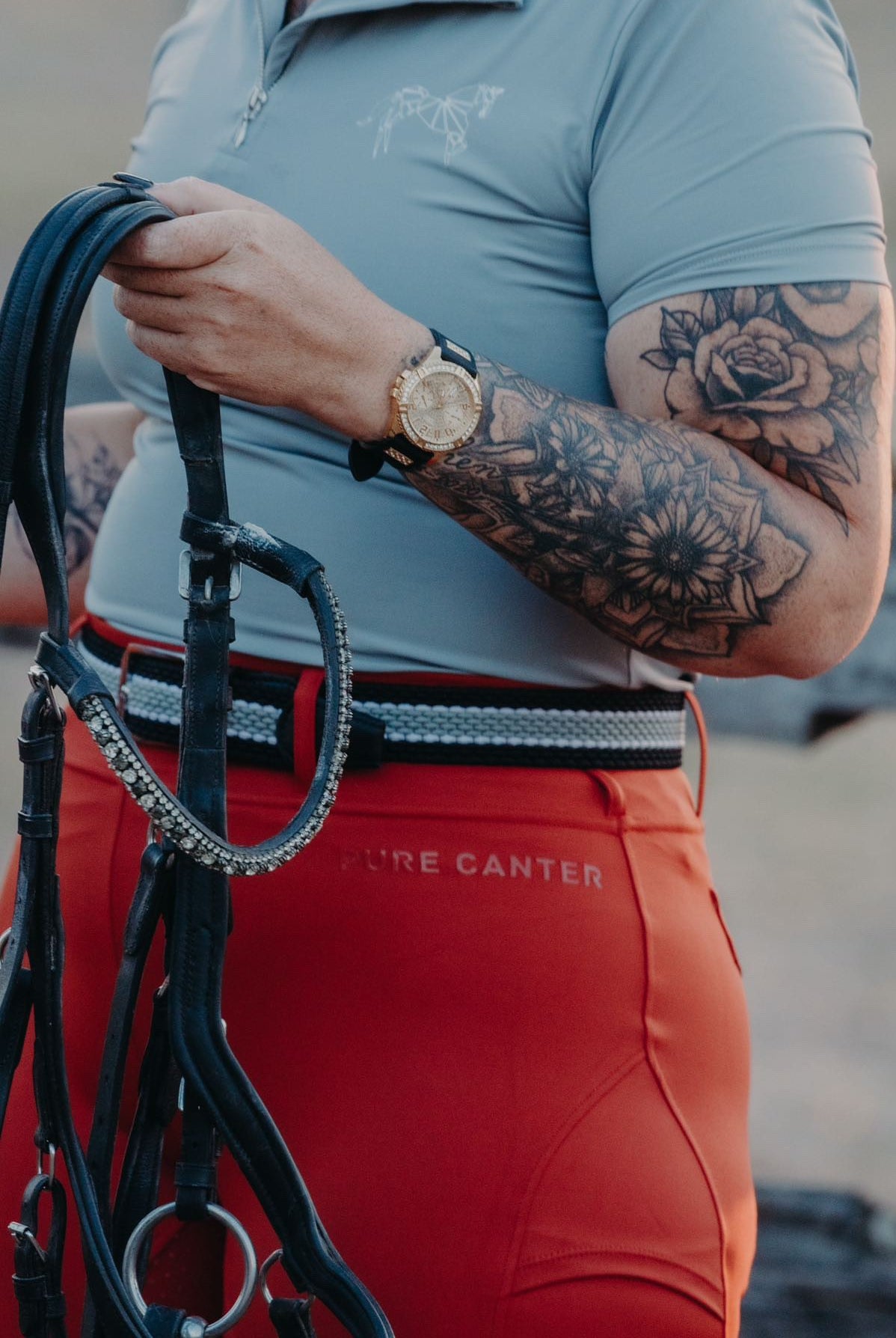 A person with tattooed arms wearing a blue shirt and bright red pants holds horse reins. The individual sports a gold watch on their left wrist. Blending equestrian fashion, the shirt features a subtle horse design while the pants, V2 Fusion Riding Tights - Outback from Pure Canter, are made of performance fabric and have "PURE CANTER" written on the hip.
