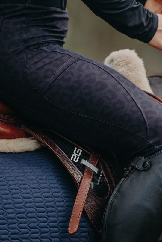 Close-up of a person riding a horse, showing the rider's lower body, saddle, and horse's back. The rider is wearing dark Pure Canter Fusion Tights - Black Leopard and black riding boots. The saddle is brown leather with a dark blue quilted saddle pad underneath.