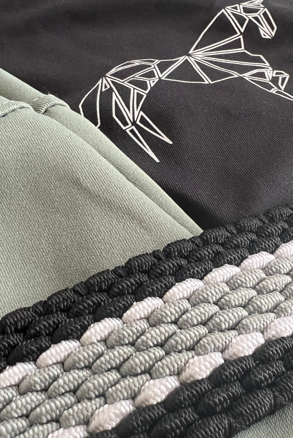 Close-up of black and green fabric with an embroidered geometric horse design on the black section. Below the fabric, Pure Canter's The Braided Belt features a braided strap with interwoven black, gray, and white strands and a secure buckle for added functionality.