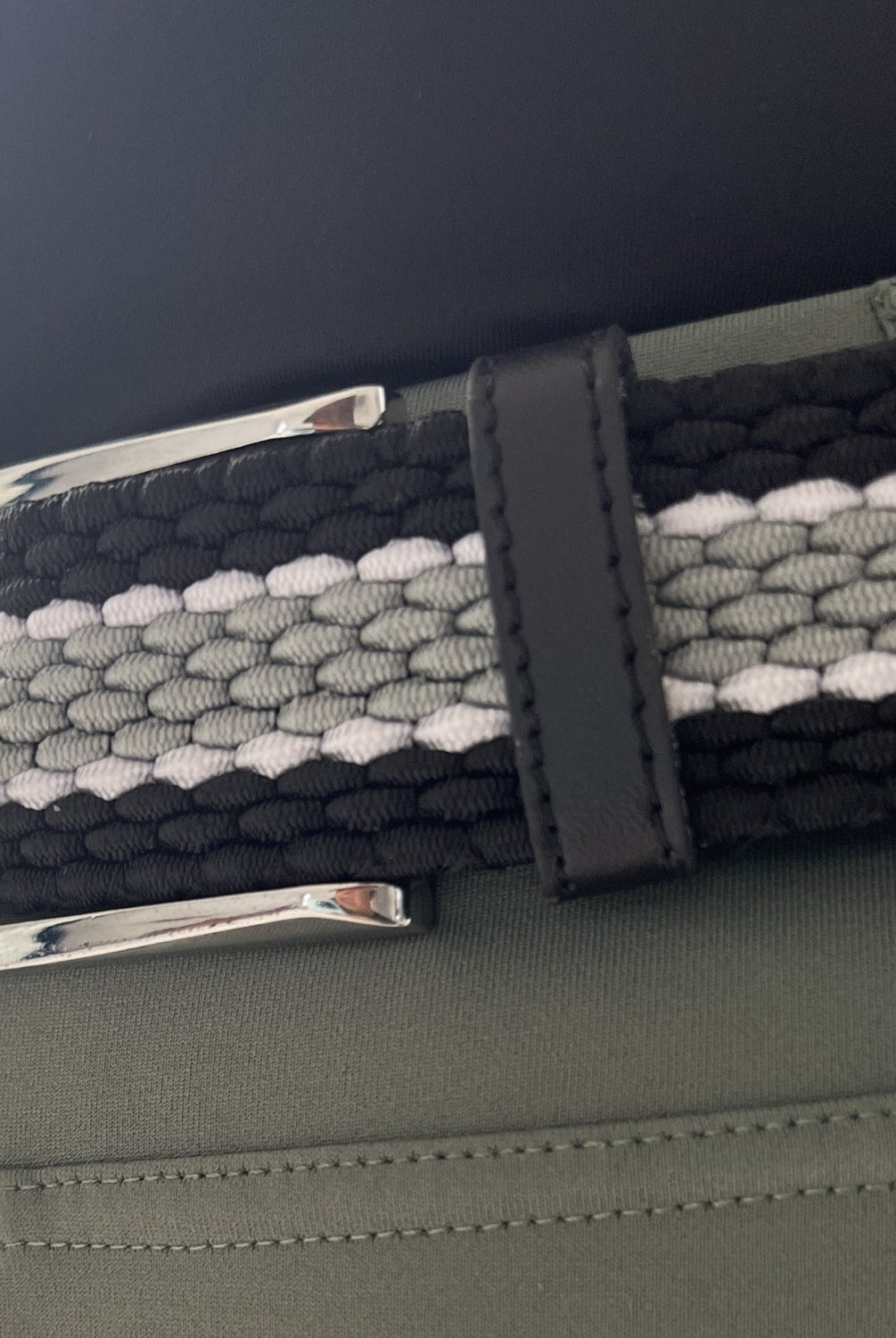 Close-up of Pure Canter's The Braided Belt worn on green pants. The belt features a secure buckle and a pattern consisting of black, gray, and white woven strands with a slight stretch. The metallic silver buckle and black leather loop add a touch of sophistication.