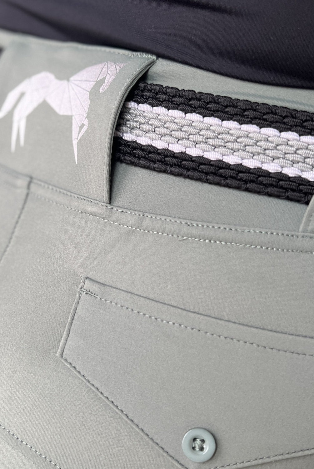 Close-up view of a person wearing light gray pants that have a buttoned back pocket, standing with their back facing the camera. The pants feature The Braided Belt from Pure Canter, showcasing a black and white geometric horse design emblem on the waistband, offering both style and a secure buckle.