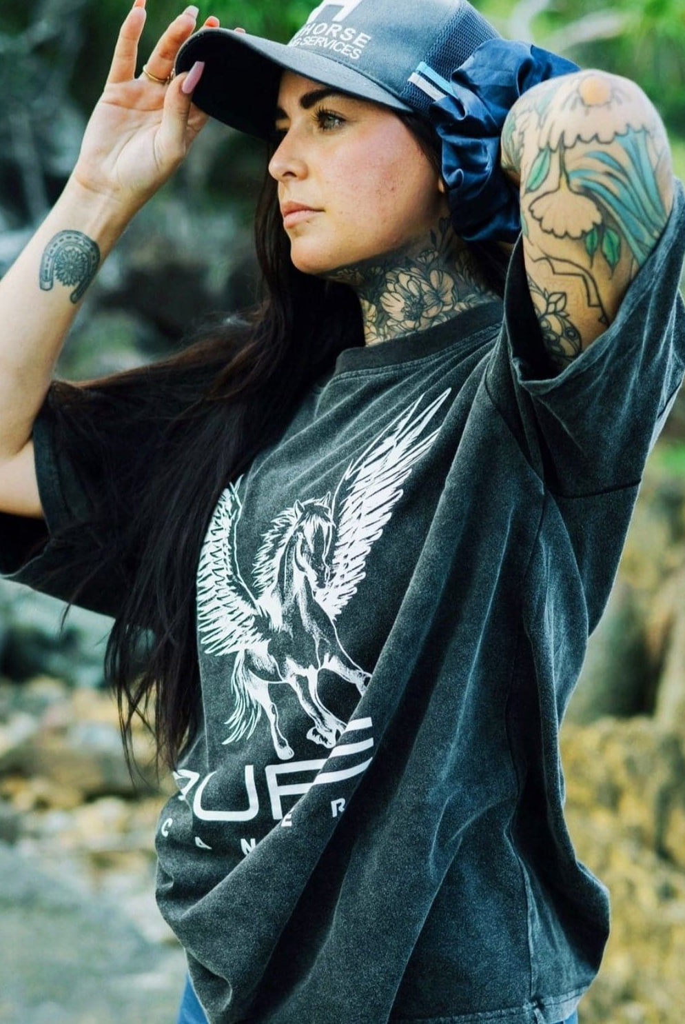 A tattooed person with long dark hair is outdoors, adjusting their hat. They are wearing a super comfy, dark baggy PURE Boyfriend Tee by Pure Canter with a stunning white Pegasus design on it. Rocks and greenery are visible in the background.