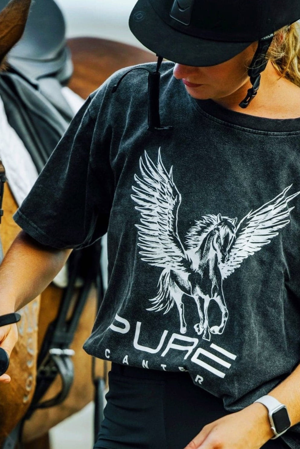 A person wearing a helmet and a baggy Pure Canter PURE Boyfriend Tee with a Pegasus design is leading a horse. The shirt has the text "PURE CANTER." The individual holds the horse's reins and looks downward while standing beside the saddled horse, exuding a relaxed, super comfy vibe.