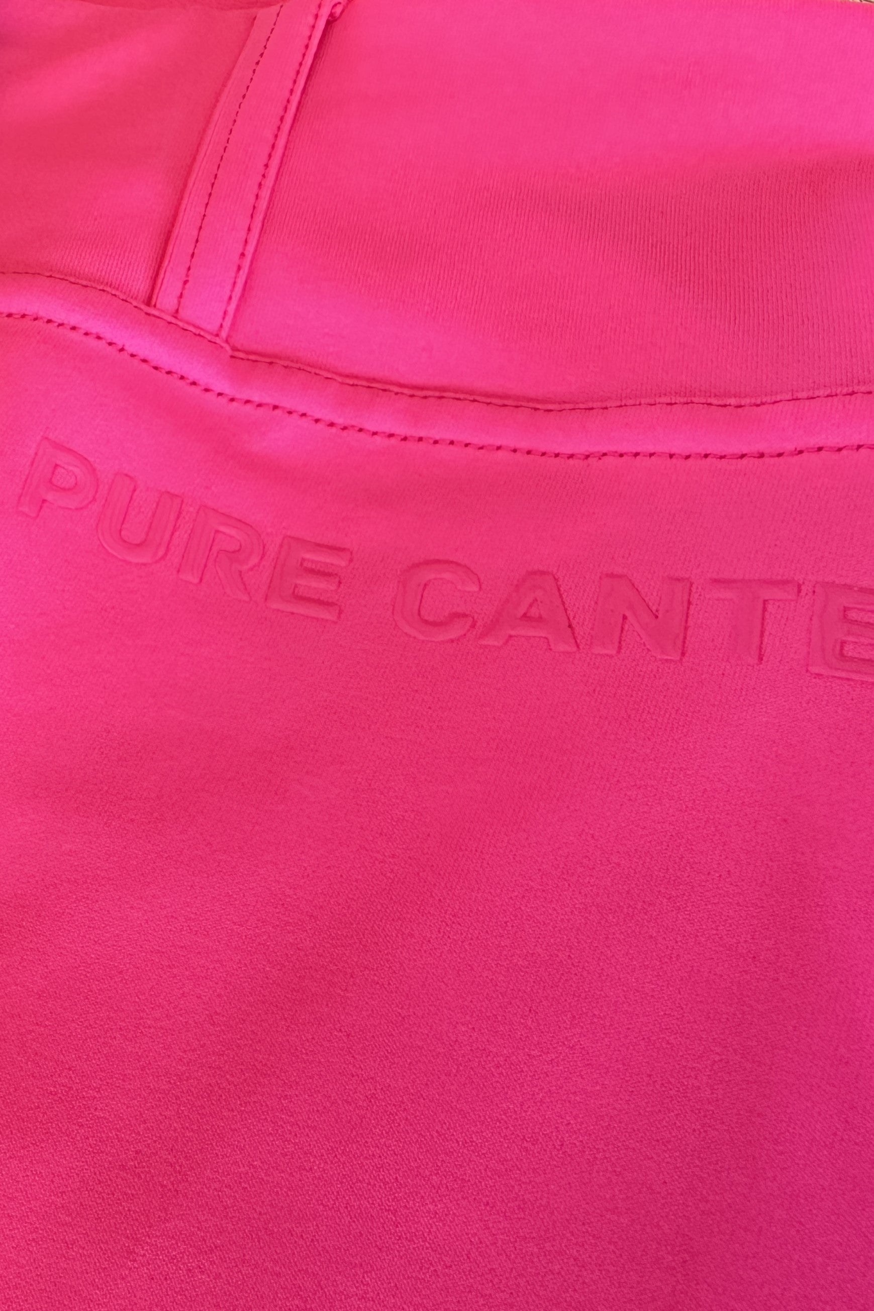 Close-up image of a bright pink performance fabric with stitching details. The words "Pure Canter" are embossed onto the fabric, appearing slightly raised and subtly visible due to the similar color, capturing the essence of equestrian fashion in the V2 Fusion Riding Tights - Hot Pink by Pure Canter.
